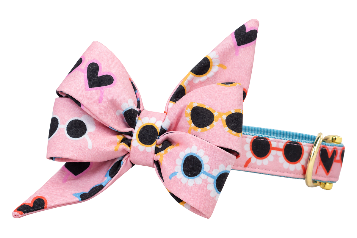 Dog collar featuring a large belle bow in Cute Shades fabric, a light pink print with black, yellow, light blue and red-orange sunglasses and black hearts, attached to a matching Cute Shades collar with a nylon inner lining and gleaming gold hardware, including a D-ring and buckle.