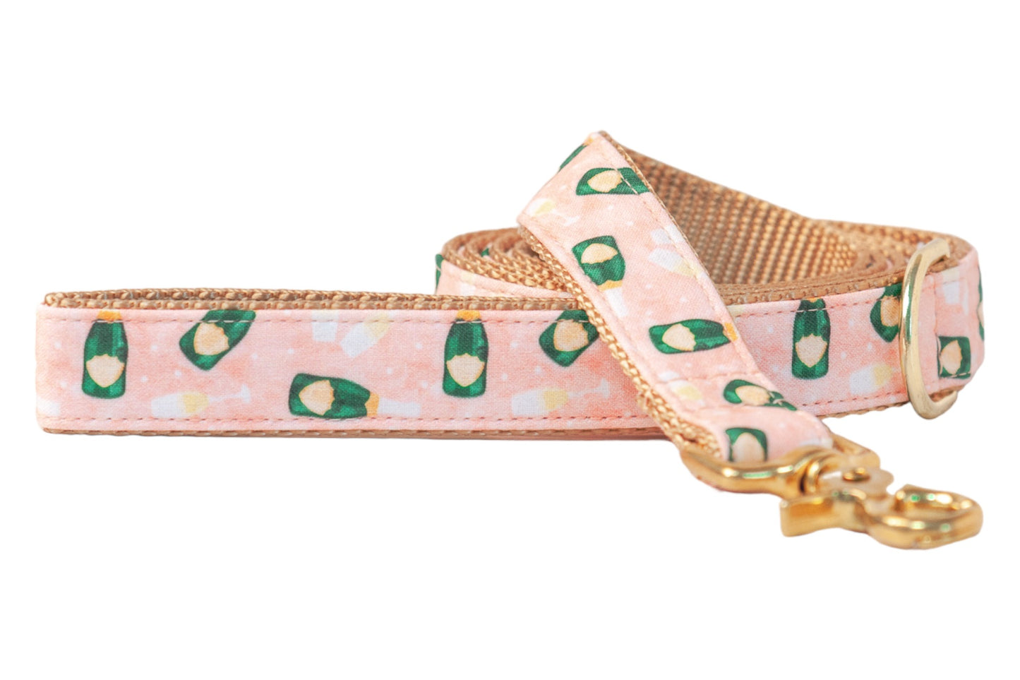 Stylish dog leash featuring a soft pink fabric adorned with repeating illustrations of champagne bottles and toasting flutes. The leash is accented with golden hardware and a tan woven underside for strength and visual texture.