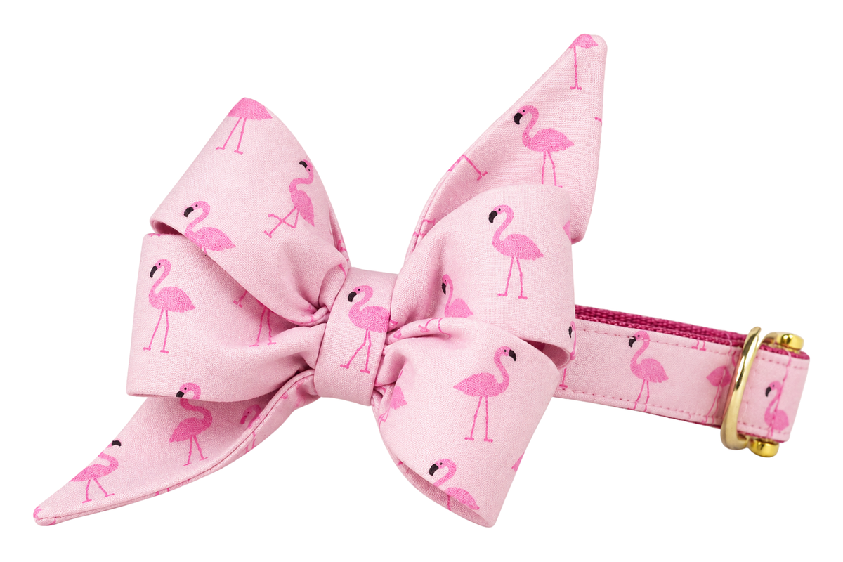 Dog collar featuring a large belle bow in Flamingo Fun fabric, a pale blush pink fabric with small pink flamingos, attached to a matching Flamingo Fun collar with a nylon inner lining and gleaming gold hardware, including a D-ring and buckle.