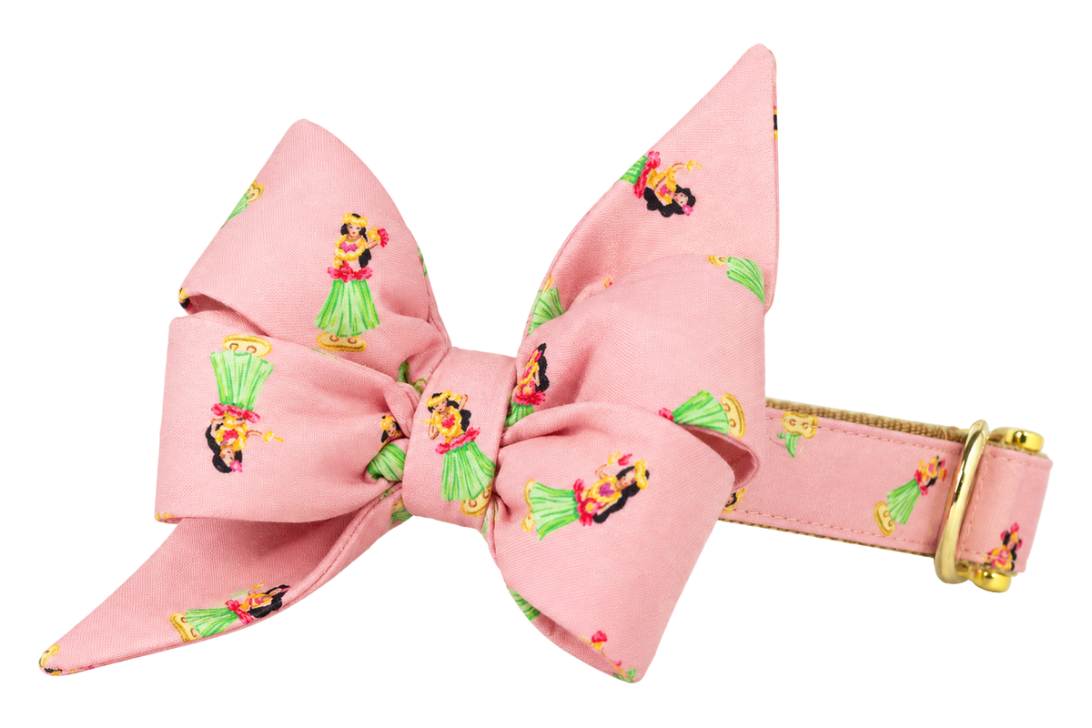 Dog collar featuring a large belle bow in Hula Girls fabric, a soft pink fabric with small hula girls in green grass skirts, attached to a matching Hula Girls collar with a nylon inner lining and gleaming gold hardware, including a D-ring and buckle.