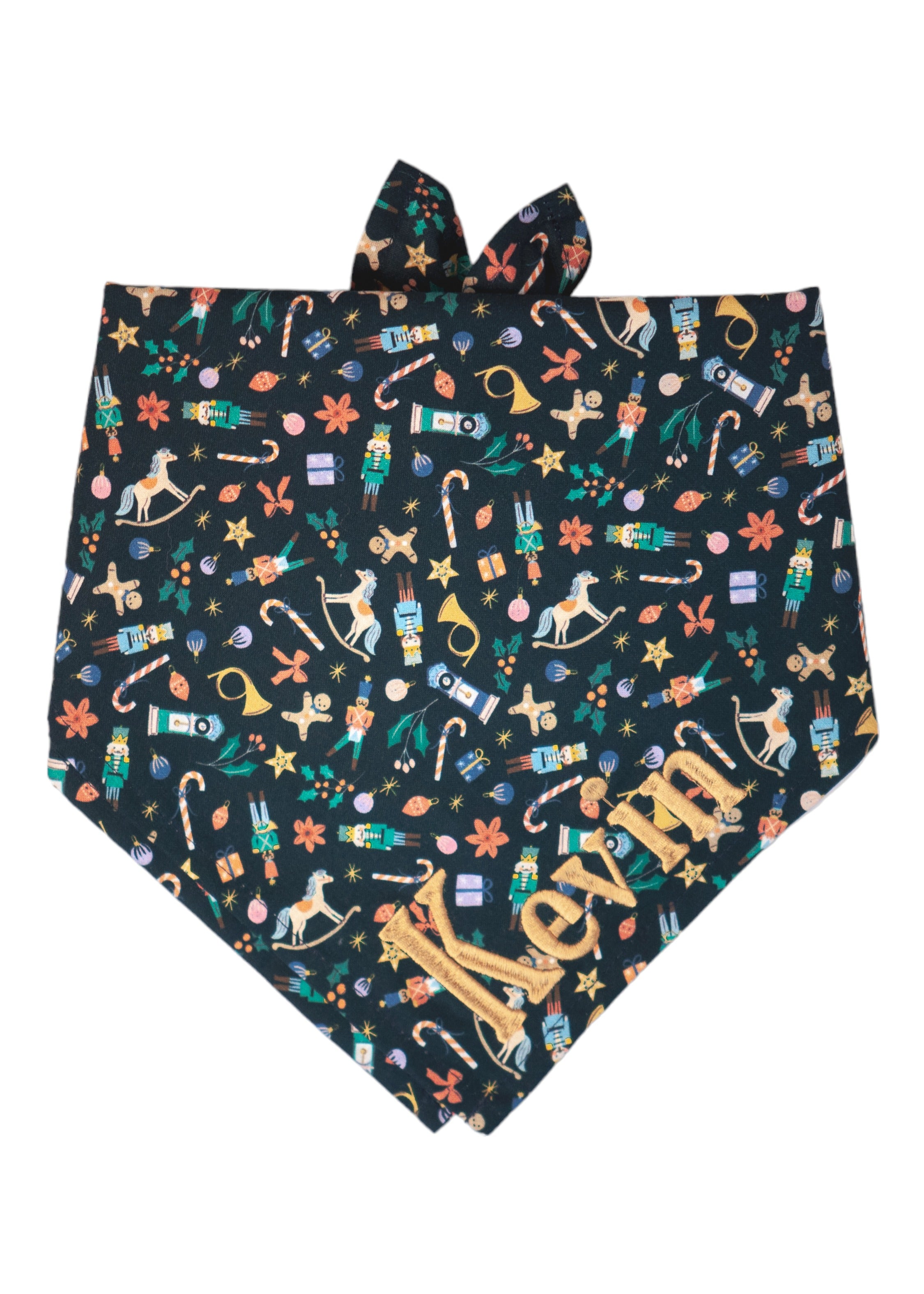 A festive dog bandana with a navy blue background, with a dense pattern of holiday icons including nutcrackers, rocking horses, candy canes, gingerbread men, stars, holly sprigs, gift boxes, French horns, and ornaments in cheerful Christmas colors. The name "Kevin" is embroidered on the bottom right.