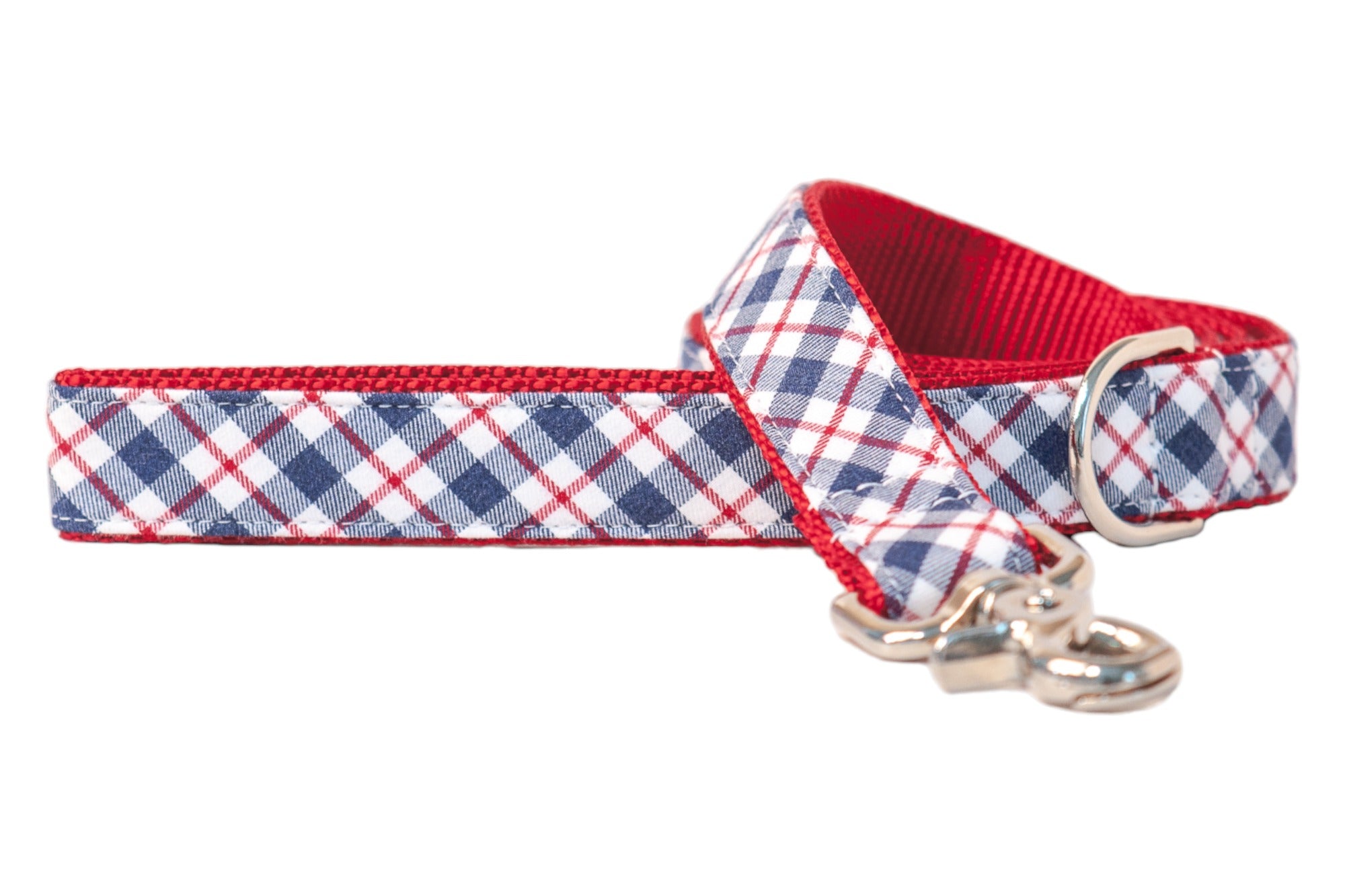 A stylish dog leash with a red, white, and navy blue crisscross plaid design. Red interior webbing adds bold contrast, while the silver metal hardware includes a swivel clip and a D-ring near the loop handle.