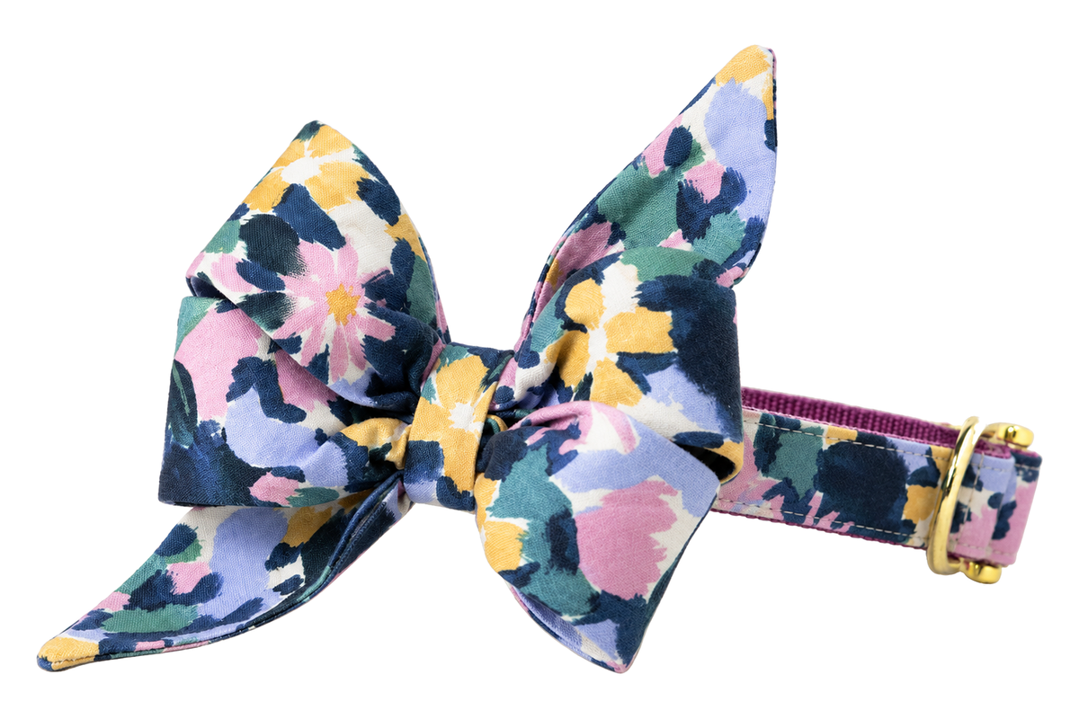 Dog collar featuring a large belle bow in Petal Pop fabric, a navy print with large painterly pink, lavender, yellow, sage and cream petals, attached to a matching Petal Pop collar with a nylon inner lining and gleaming gold hardware, including a D-ring and buckle.