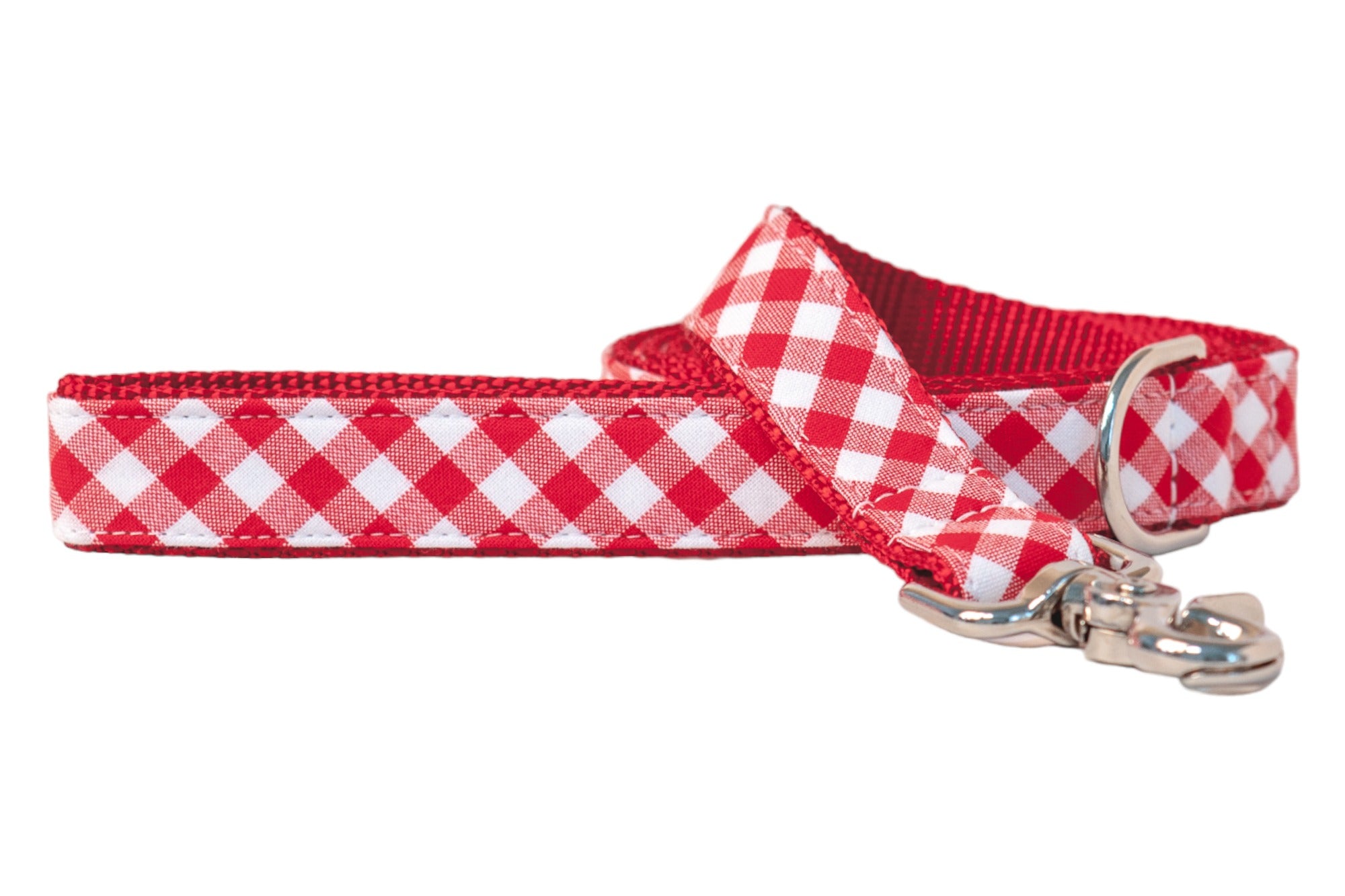 High-detail view of a red checkered dog leash, combining a traditional plaid fabric overlay with a sturdy red nylon webbing backing. The leash features tight topstitching for durability, a metal swivel clip for smooth leash movement, and a D-ring loop at the handle for hanging or attaching small items. The vibrant red webbing adds both structural support and visual dimension.
