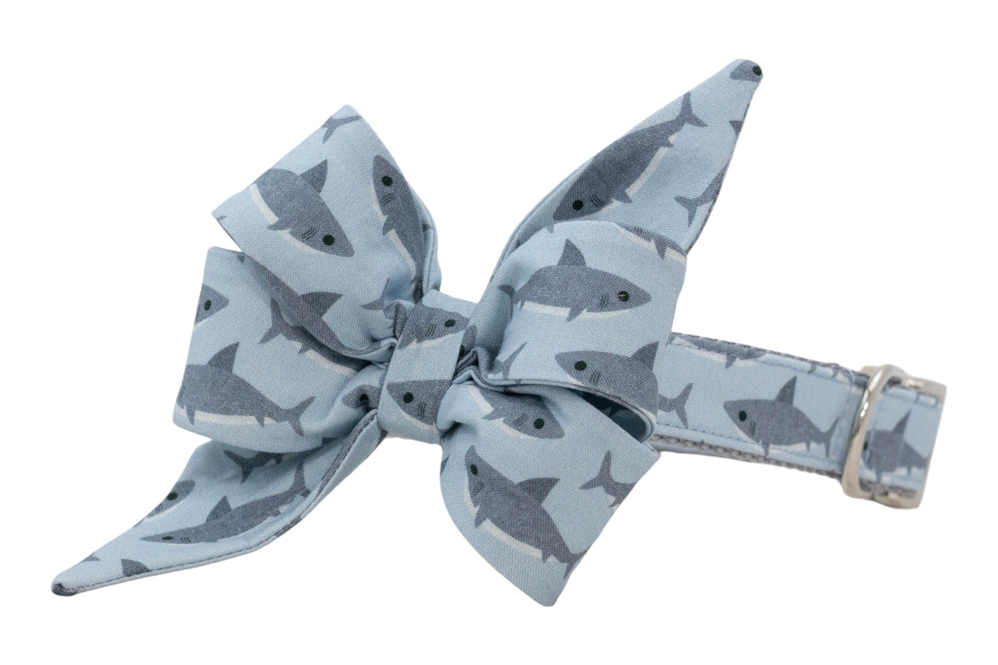 Pale blue pet collar with a tied bow detail, decorated with small dark gray sharks and finished with a metal D-ring.