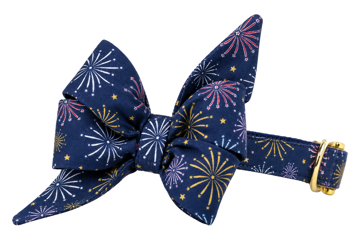 Dog collar featuring a large belle bow in Sparks Fly fabric, a deep navy blue fabric with fireworks bursts in gold, pink, lavender and light blue and small gold stars, attached to a matching Sparks Fly collar with a nylon inner lining and gleaming gold hardware, including a D-ring and buckle.