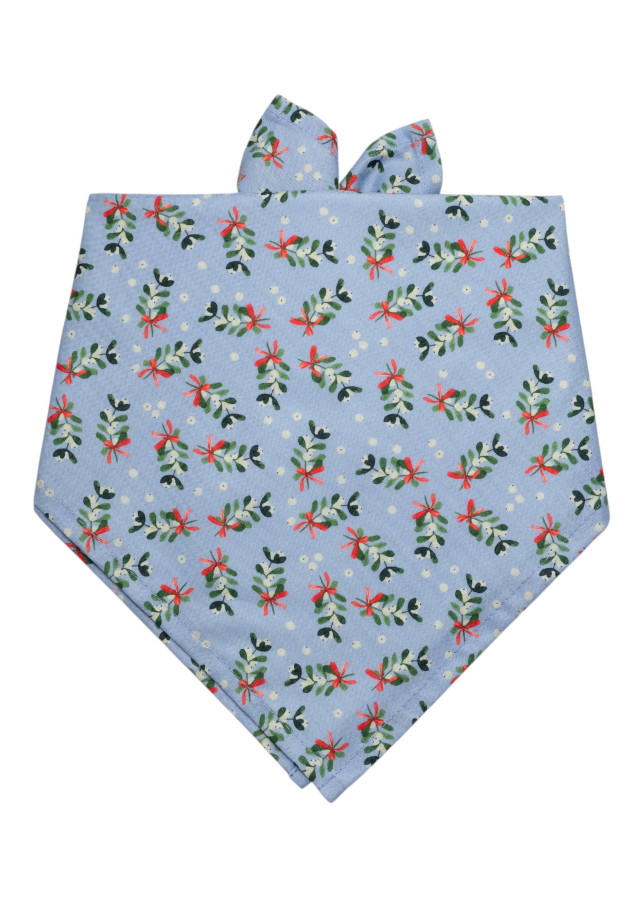 A festive dog bandana made from light blue fabric with a repeating pattern of mistletoe sprigs, berries, and ribbon bows. The design has neatly stitched edges and a fold-over top for easy tying around a dog's neck, offering a soft and wintry holiday look.