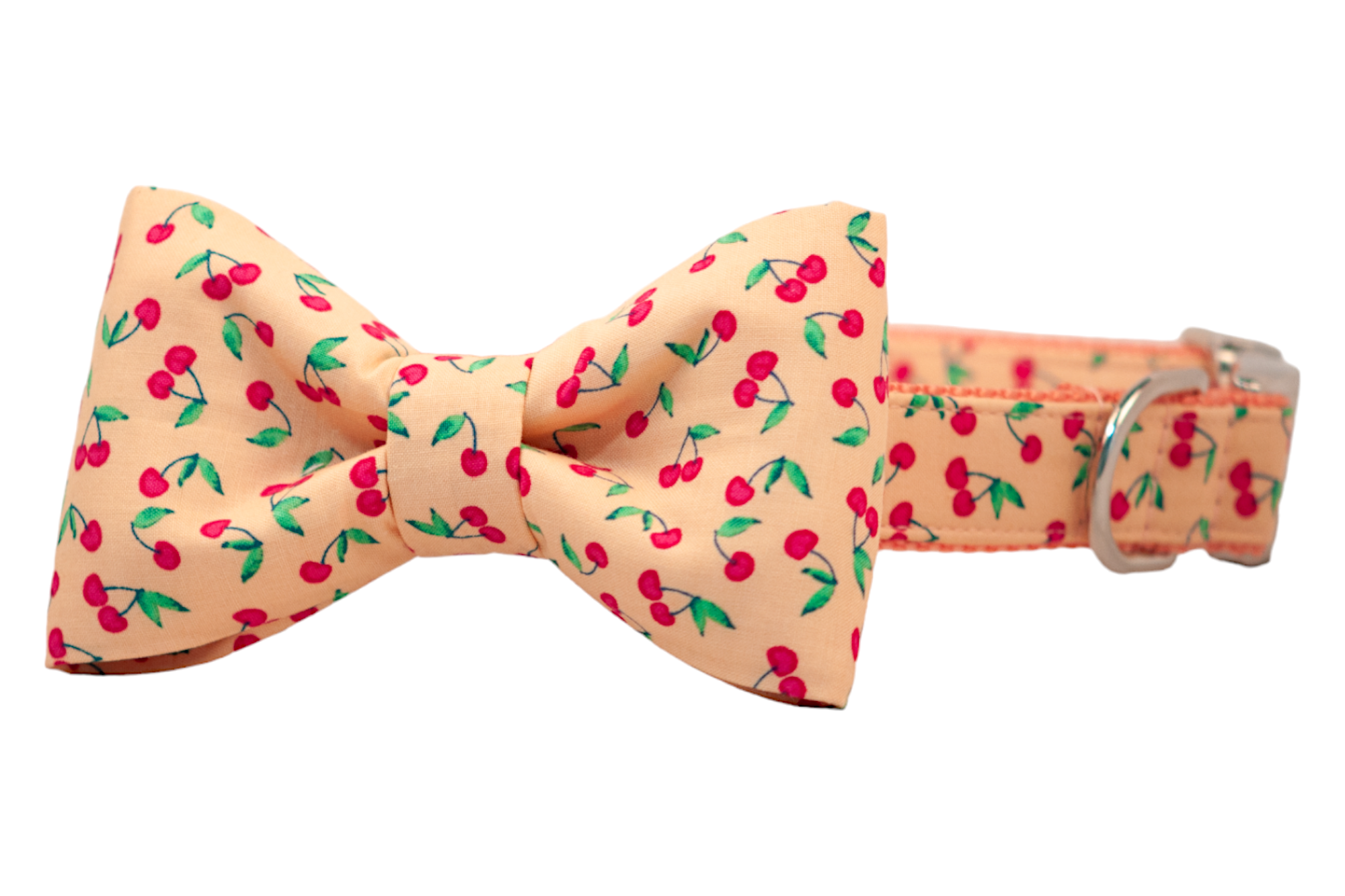 A cheerful fabric bow tie collar for dogs, printed with clusters of red cherries and green leaves on a soft peach background. The bow is center-cinched for a classic shape, and the collar is finished with a shiny silver D-ring and buckle.