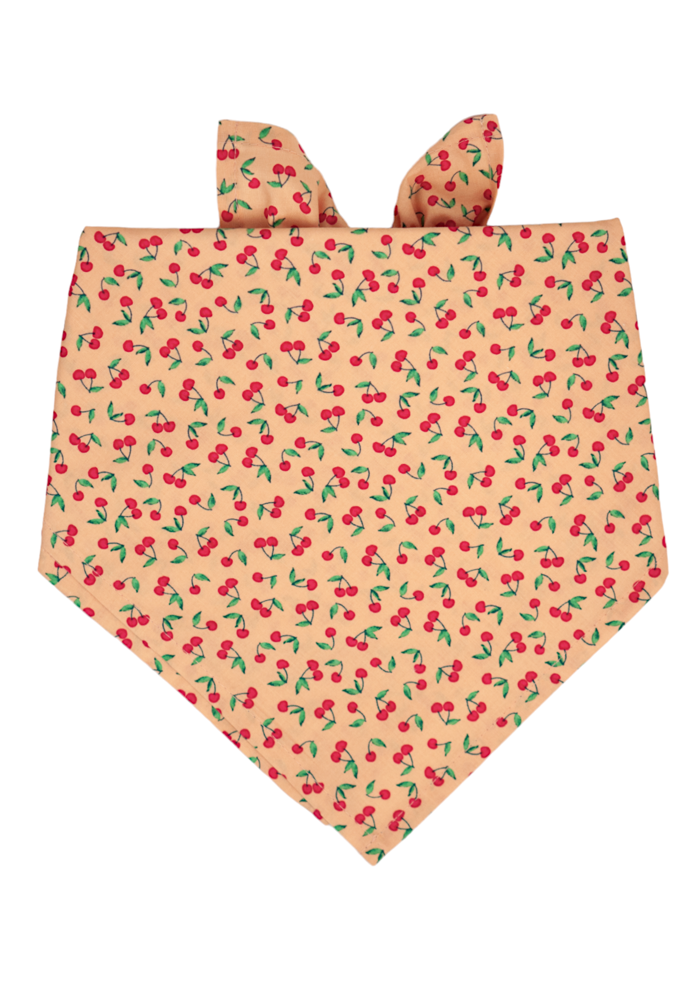 A soft peach-colored dog bandana featuring a playful cherry print with small red cherries and bright green leaves. The bandana is folded in a classic triangle shape with neatly stitched hems and visible tie points at the top.