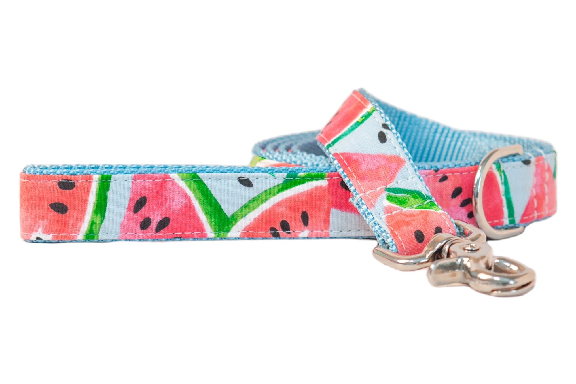 A dog leash with a bright watermelon print on light blue fabric, featuring red-pink fruit sections, green-striped rinds, and scattered black seeds. The underside is made of sky-blue woven nylon webbing, and the leash is finished with silver-tone hardware, including a swivel snap hook and a D-ring.