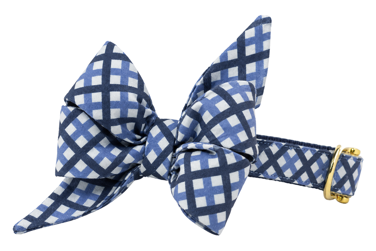 Dog collar featuring a large belle bow in Windowpane Plaid fabric, a navy and periwinkle blue lattice check on white, attached to a matching Windowpane Plaid collar with a nylon inner lining and gleaming gold hardware, including a D-ring and buckle.