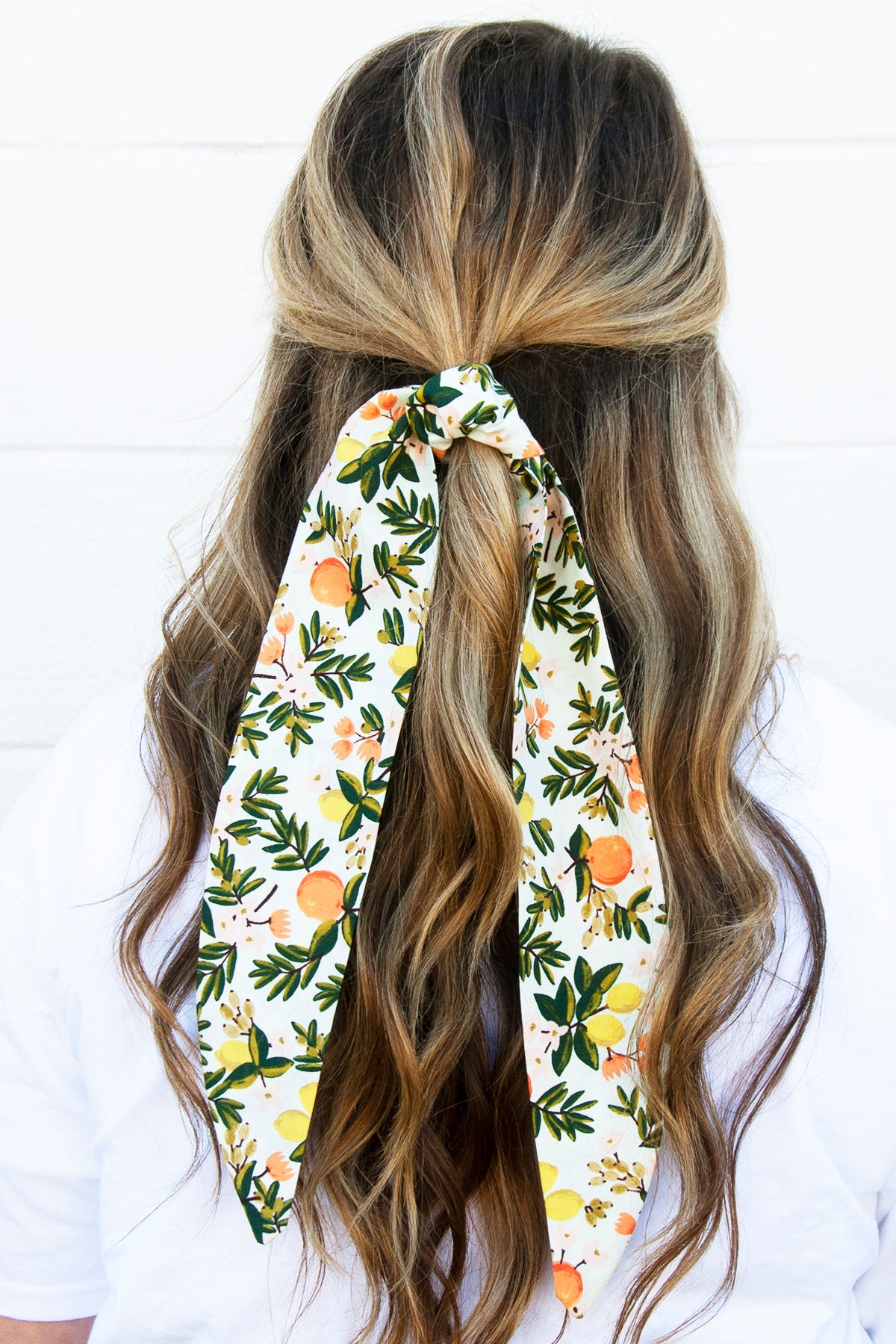 Back view of a woman with loose waves wearing a statement hair scarf in a cheerful citrus and botanical pattern, sunny, playful, and ready for summer vibes.