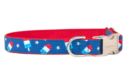A detailed view of a patriotic dog collar  showcasing a blue fabric design covered in white stars and vibrant red, white, and blue popsicles. The collar has an inner red webbing, fastens with a metal clasp, and includes a shiny silver D-ring for functional use.