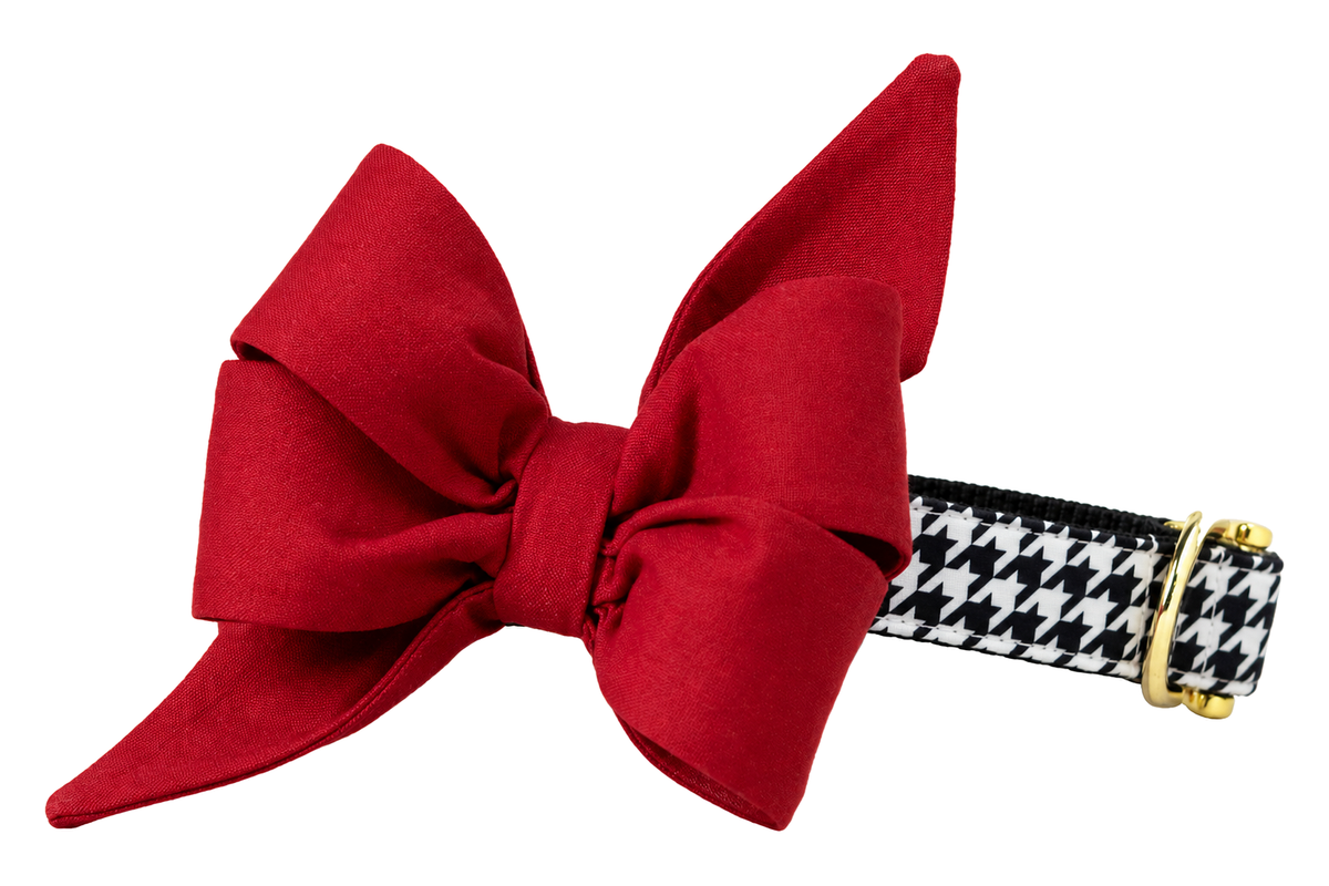 Dog collar featuring a large belle bow in Alabama On Houndstooth fabric, a solid crimson bow paired with a black and white houndstooth, attached to a matching Alabama On Houndstooth collar with a nylon inner lining and gleaming gold hardware, including a D-ring and buckle.