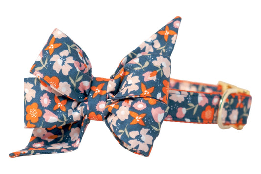 Dog collar adorned with an oversized four-loop belle bow in a vibrant floral print of blossoms with leafy stems on a dark blue background. The belle bow’s top loops are elongated and pointed, while the bottom loops are fuller and angled outward, giving it a feminine style. The matching collar is adjustable with metal hardware.