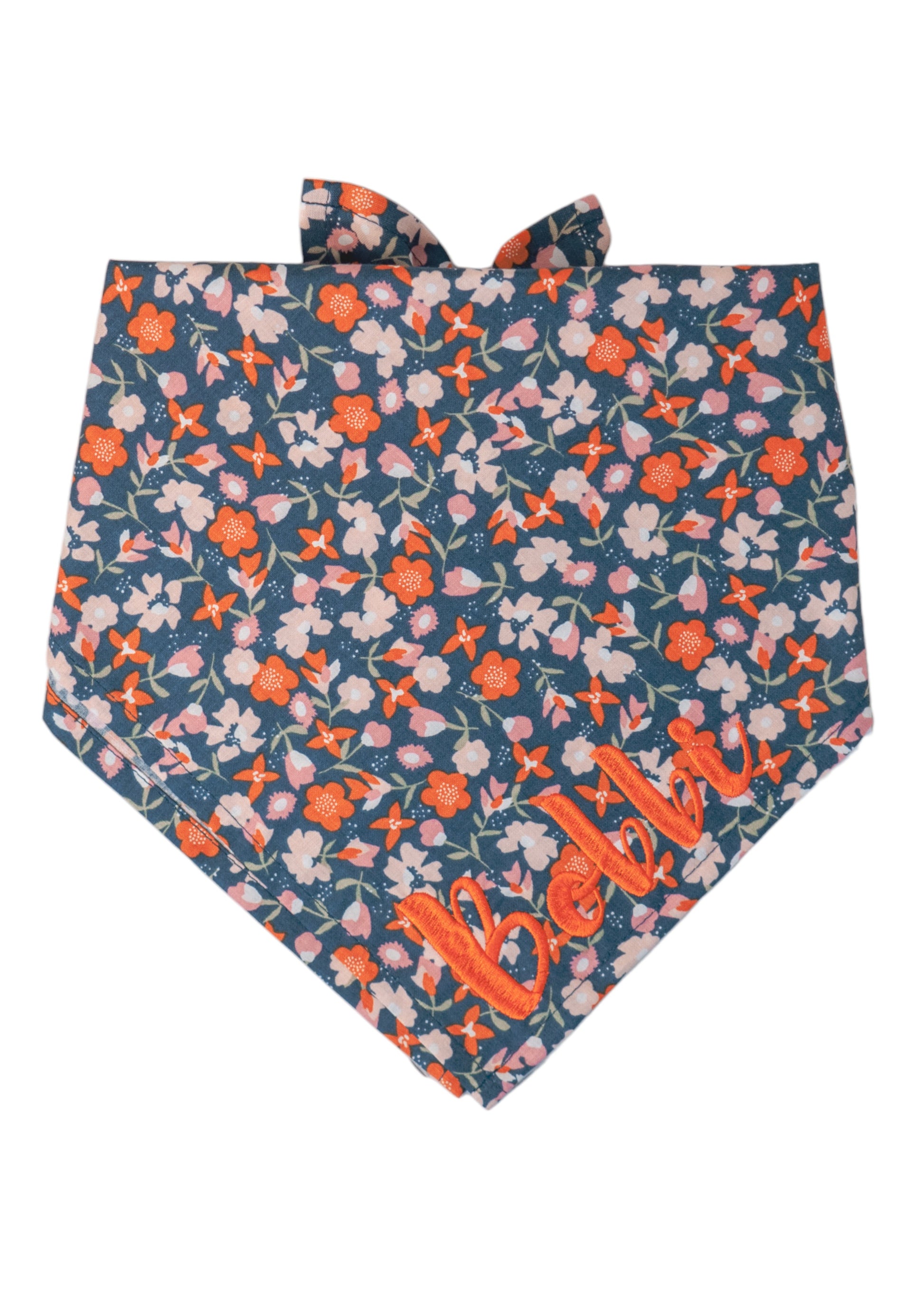 Dog bandana made from smooth cotton fabric with a colorful flower pattern featuring blooms on a dark blue base. The bandana has the name "Bobbi" embroidered on the right side, is neatly hemmed, folded , and is designed to be tied at the top for securing around your pup's neck.