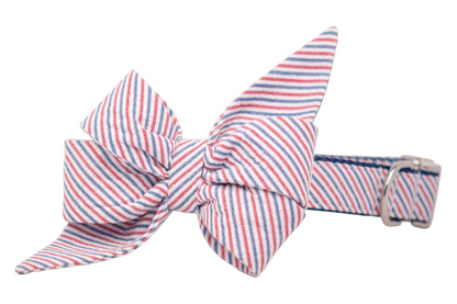 A stylish dog collar featuring a large, oversized and feminine belle bow made from seersucker with diagonal red, white, and blue pinstripes. The belle bow is symmetrically tied and securely attached to the collar, which is made from the same striped fabric. The collar includes a shiny silver metal buckle for fastening.