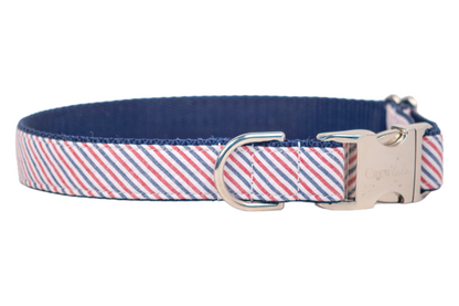 Close-up of a dog collar wrapped in a patriotic-themed seersucker fabric with thin diagonal red and blue stripes on a white background. The collar includes a silver D-ring for leash attachment and a polished metal clasp engraved with branding. The interior of the collar is a dark navy webbing.