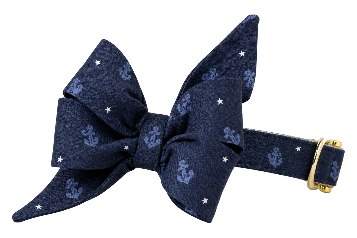 Dog collar featuring a large belle bow in Anchors Away fabric, a navy blue fabric with small tonal periwinkle blue anchors and tiny white stars, attached to a matching Anchors Away collar with a nylon inner lining and gleaming gold hardware, including a D-ring and buckle.