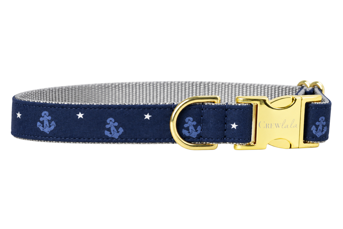 Anchors Away dog collar made with a navy blue fabric with small tonal periwinkle blue anchors and tiny white stars, lined with light gray nylon webbing and finished with gold hardware, including a D-ring and a Crewlala logo buckle.