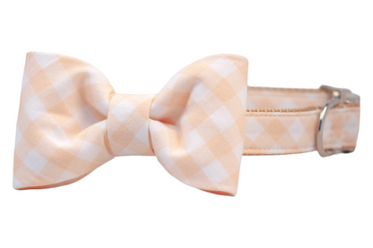 A charming dog collar featuring a soft apricot and white checkered bow tie, neatly centered and attached to a matching fabric-wrapped collar with tan webbing and polished silver hardware, perfect for special occasions or everyday cuteness.