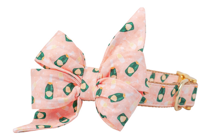A large decorative dog belle bow attached to a dog collar, both have blush pink fabric patterned with miniature green champagne bottles and filled glasses. The bow features dramatic, pointed tails and full, structured loops, creating a statement look. The collar includes gold hardware and reinforced stitching, blending style with durability.