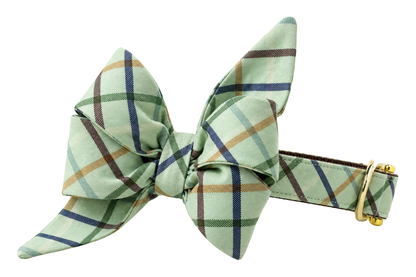 Dog collar featuring a large belle bow in Big Bear Plaid fabric, a pale sage green fabric with a windowpane plaid of navy, dark green and tan lines, attached to a matching Big Bear Plaid collar with a nylon inner lining and gleaming gold hardware, including a D-ring and buckle.