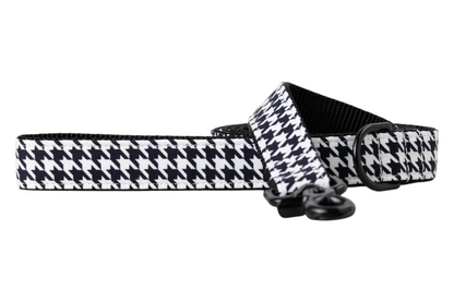 A dog leash with a black and white houndstooth fabric overlay, backed with sturdy black nylon webbing for durability. The leash is shown partially coiled and includes matte black hardware, including a swivel clasp and D-ring for secure attachment and accessory clips. A modern, stylish accessory for fashionable pets.