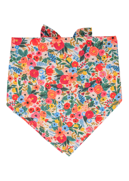 Close-up of a dog bandana with a festive floral motif. The fabric is covered in an intricate mix of bright red, orange, pink, yellow, and blue flowers, surrounded by green leafy stems and tiny scattered berries, all printed on a pale background. Two tie ends are visible at the top for securing around a pet’s neck.