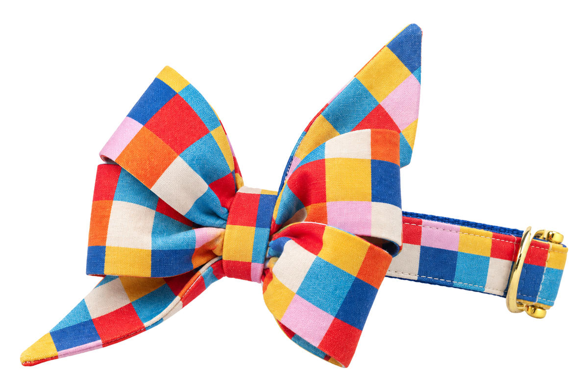 Dog collar featuring a large belle bow in Block Party fabric, a bold colorblock check of red, cobalt blue, yellow, orange, pink and cream squares, attached to a matching Block Party collar with a nylon inner lining and gleaming gold hardware, including a D-ring and buckle.