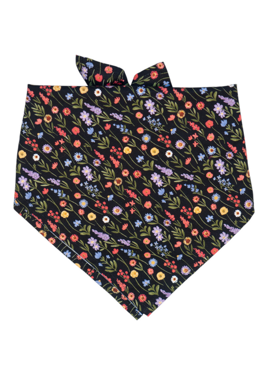 Black bandana folded in a triangle shape, featuring an all-over print of flowers in shades of pink, yellow, and coral, purple with green foliage. Two ear-like knots are tied at the top.