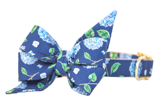 Navy floral dog collar with a statement belle bow attached, made from soft cotton fabric. The belle bow has a sculpted, elegant shape and a botanical hydrangea design in blue. The collar features a metal D-ring and buckle for fastening.