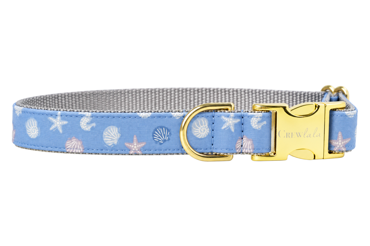 Blue Sea Shells dog collar made with a periwinkle blue fabric with small white seashells and starfish, lined with light gray nylon webbing and finished with gold hardware, including a D-ring and a Crewlala logo buckle.