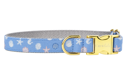 Blue Sea Shells dog collar made with a periwinkle blue fabric with small white seashells and starfish, lined with light gray nylon webbing and finished with gold hardware, including a D-ring and a Crewlala logo buckle.