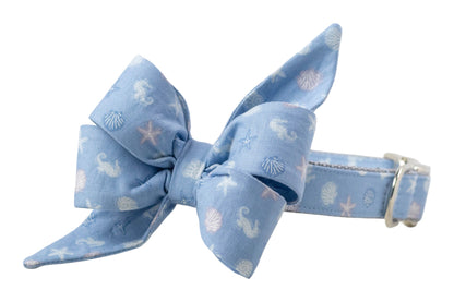 Pale blue pet collar with a tied bow detail, decorated with small shells, seahorses, and starfish in a subtle coastal print.