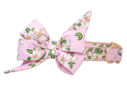 Dog collar featuring a romantic pink fabric printed with white magnolia blossoms and leaves. The collar is adorned with a large, structured belle bow made of the same floral fabric, with dramatic pointed ends and layered folds. The collar is reinforced with nylon webbing and finished with a metal buckle and D-ring for a polished, feminine look.