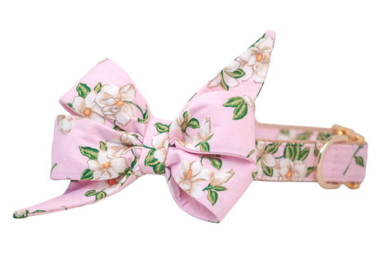 Dog collar featuring a romantic pink fabric printed with white magnolia blossoms and leaves. The collar is adorned with a large, structured belle bow made of the same floral fabric, with dramatic pointed ends and layered folds. The collar is reinforced with nylon webbing and finished with a metal buckle and D-ring for a polished, feminine look.