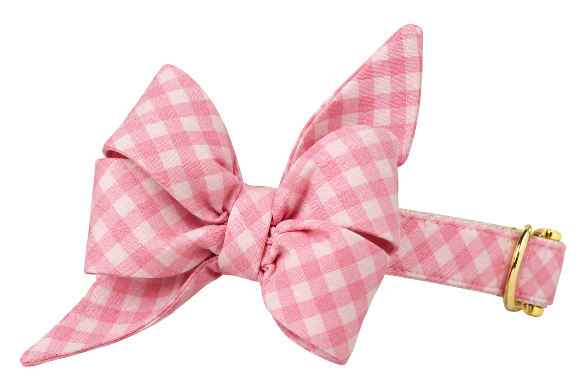 Dog collar featuring a large belle bow in Blush and Bashful Picnic Plaid fabric, a soft blush pink and white gingham check, attached to a matching Blush and Bashful Picnic Plaid collar with a nylon inner lining and gleaming gold hardware, including a D-ring and buckle.