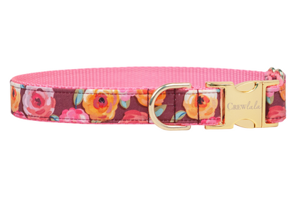 A fabric dog collar with a bold floral print featuring roses in warm fall tones on a burgundy background. The collar is lined with nylon webbing and includes a shiny metal buckle engraved with the brand name "Crewlala" and a matching D-ring.