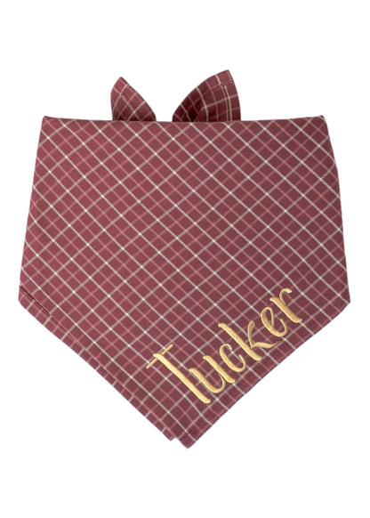 Dog bandana in burgundy fabric with a soft plaid pattern, personalized with the name “Tucker” embroidered near the point. The bandana features two tie-up flaps at the top for secure wear.