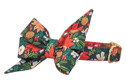 A festive dog collar featuring a large, double-layered belle bow with long, pointed tails. The fabric has a dark green background and is decorated with a vibrant holiday print including poinsettias, pinecones, flowers, blossoms, holly berries, and green foliage. The collar is adjustable and fastens with a polished buckle.