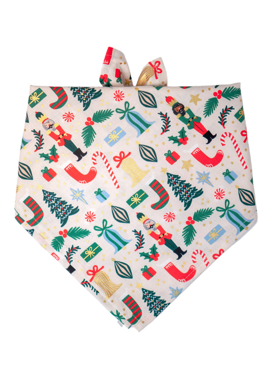 Festive dog bandana in a cream tone with a playful and colorful holiday pattern. Illustrated details include nutcrackers in red and green outfits, candy canes, stars, ornaments, wrapped presents, and Christmas trees, with metallic dots adding sparkle across the fabric.