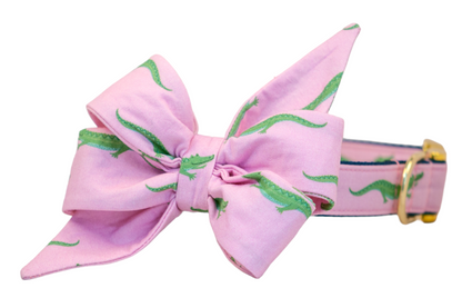 Preppy-style dog collar with an attached matching feminine belle bow in pastel pink, covered in a green crocodile pattern. The bow is elegantly tied and the collar is accented with gold metal hardware and dark blue trim for contrast.