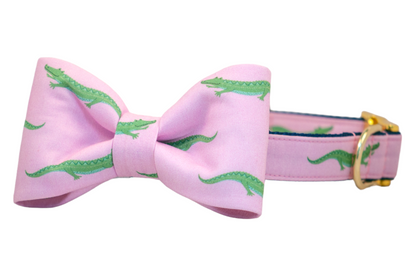 Close-up of a preppy-style dog collar featuring a structured bow tie in soft pink fabric decorated with playful green crocodiles. The collar has a gold D-ring and stitching detail along the edges, creating a polished, Southern-inspired look.
