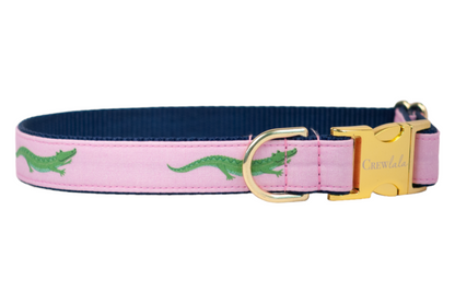 Dog collar featuring a repeating green crocodile motif on a pastel pink fabric background. The collar has a navy blue inner webbing, a gold side-release buckle engraved with “Crew LaLa,” and a gold-tone metal D-ring for leash attachment.