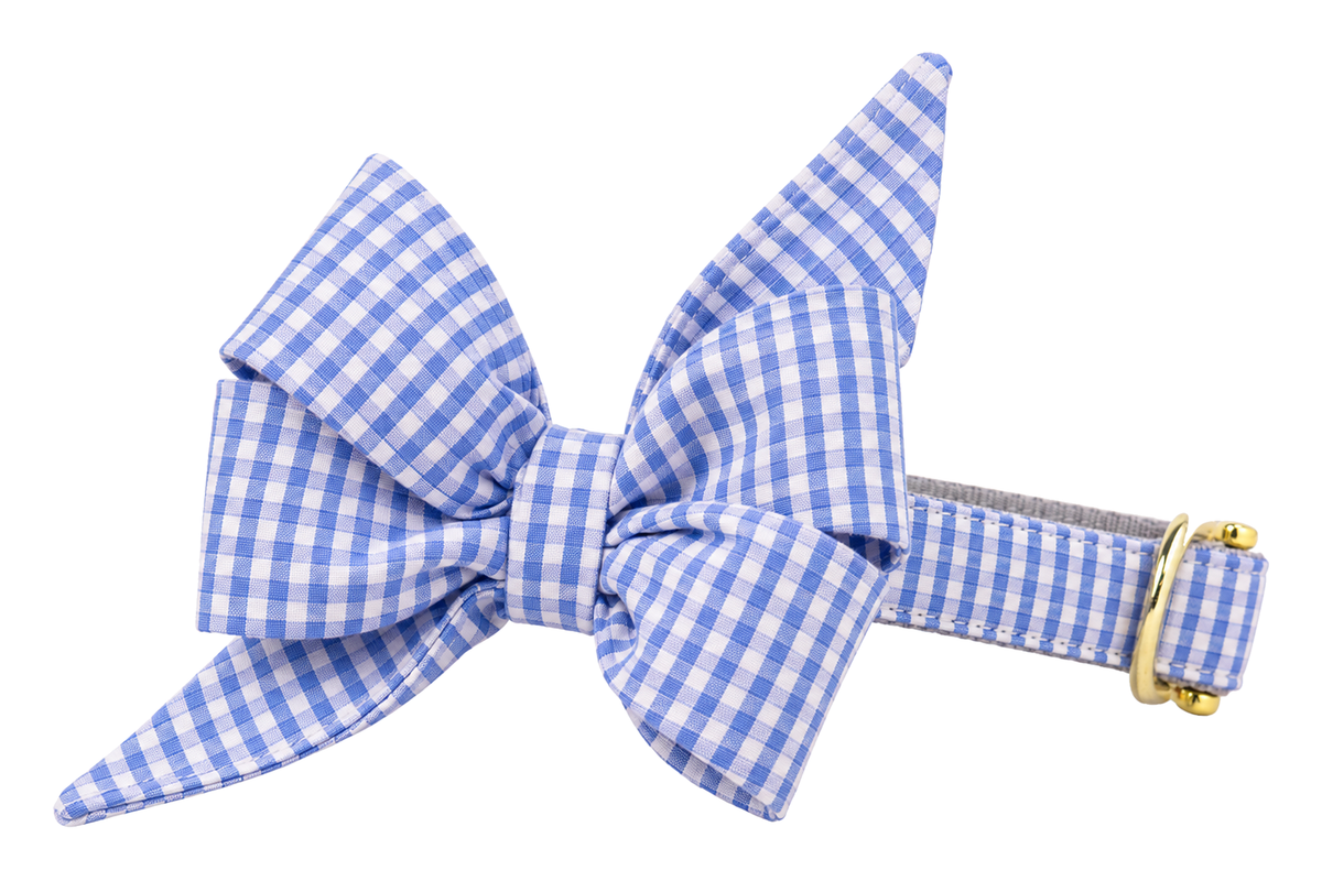 Dog collar featuring a large belle bow in Coastal Gingham fabric, a periwinkle blue and white seersucker gingham check, attached to a matching Coastal Gingham collar with a nylon inner lining and gleaming gold hardware, including a D-ring and buckle.