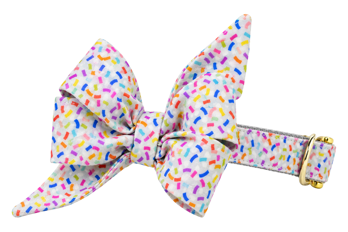 Dog collar featuring a large belle bow in Confetti Pop fabric, an off-white print with colorful confetti sprinkles in pink, purple, blue, yellow and orange, attached to a matching Confetti Pop collar with a nylon inner lining and gleaming gold hardware, including a D-ring and buckle.