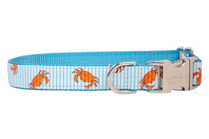 Close-up of a nautical-themed dog collar decorated with playful orange crab illustrations on a striped blue and white ribbon. The collar is reinforced with blue webbing and includes shiny silver hardware, including a clasp with the "Crew LaLa" logo and a sturdy D-ring.