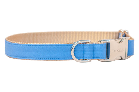 A dog collar with solid royal blue fabric stitched over tan woven nylon. It features a shiny silver metal D-ring for leash attachment and a silver side-release buckle engraved with the brand name “Crewlala.”