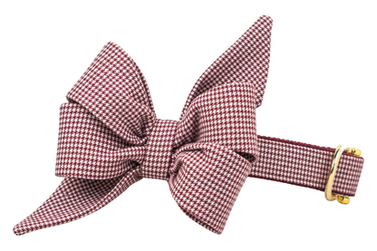 Dog collar featuring a large belle bow in Crimson Houndstooth fabric, a fine burgundy and white houndstooth check, attached to a matching Crimson Houndstooth collar with a nylon inner lining and gleaming gold hardware, including a D-ring and buckle.